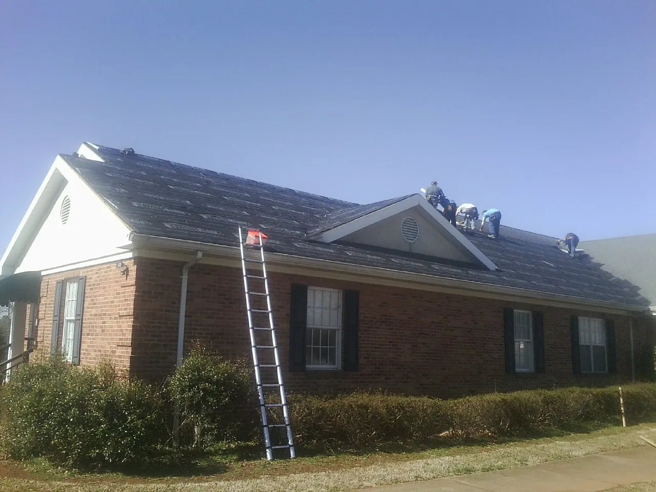 Crew tearing shingles off a brick ranch roof with a ladder set — Metal Roof Repair work by our crew in Port Barre