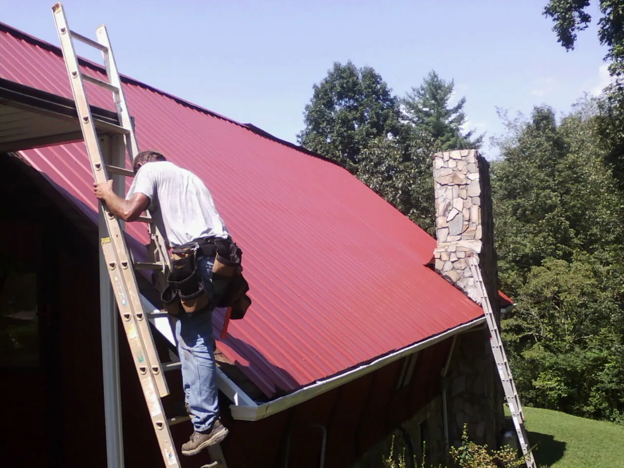 Roofer on a ladder beside a red metal roof and stone chimney — Soffit & Fascia Repair by Roofing Brothers in Port Barre